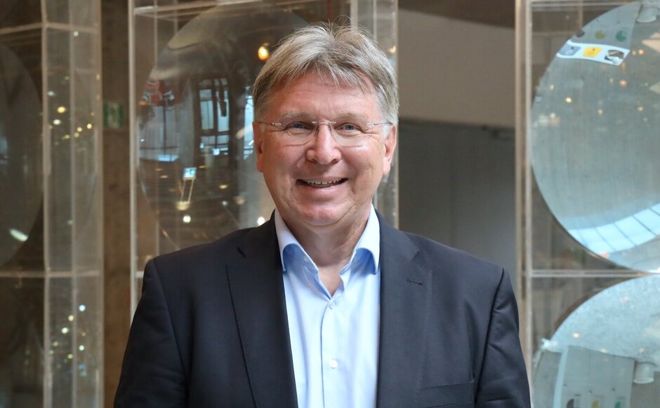 Porträtfoto: lächelnder Mann mit kurzen Haaren, Brille, grauem Sakko und hellblauem Hemd vor neutralem Hintergrund