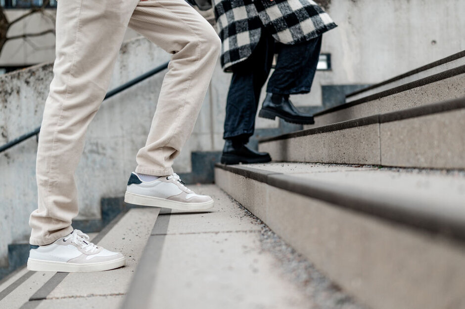 Ein Beinpaar in beigen Hosen und weißen Turnschuhen und ein Beinpaar in schwarzen Hosen und schwarzen Schnürschuhen gehen eine Betontreppe nach oben.