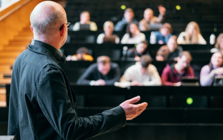 Gestikulierender Mann mit Glatze von hinten, blick auf mehrere Stuhlreihen eines Auditoriums. Diese sind voll besetzt, die Personen sind nur verschwommen zu sehen.