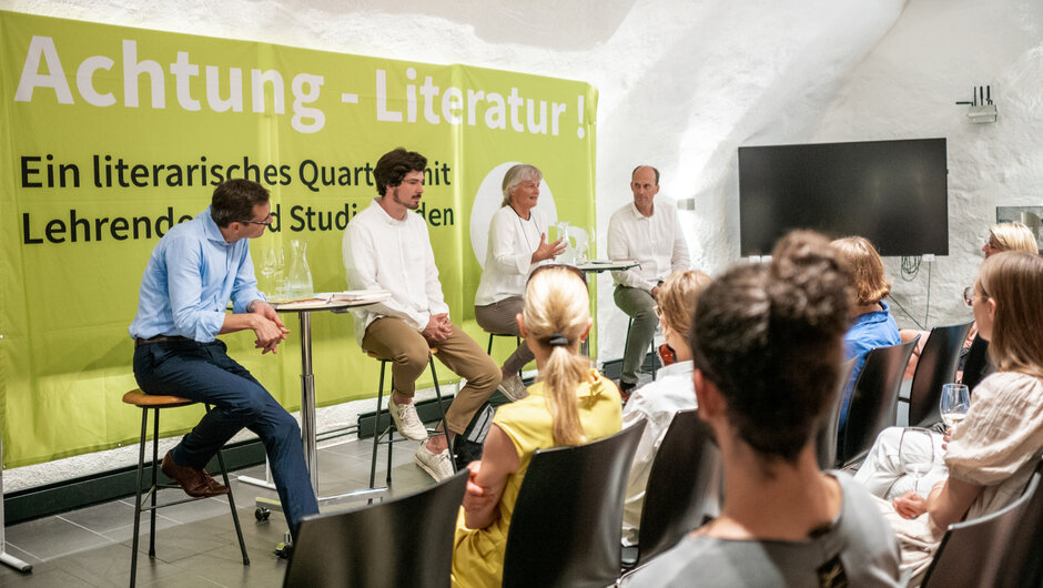 Vier Personen sitzen auf Barhockern an Stehtischen. Eine von ihnen, eine Frau mit grauen Haaren, spricht gestikulieren, die anderen hören aufmerksam zu. Im Hintergrund steht ein Banner mit der Aufschrift "Achtung - Literatur!". (öffnet Vergrößerung des Bildes)