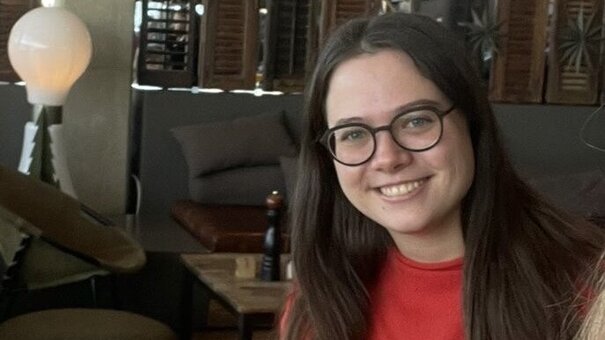 Katharina Reitzer, eine braunhaarige junge Frau, lächelt in die Kamera. Sie sitzt in einem öffentlichen Raum mit mehreren Sofas und Couchtischen sowie einer Stehlampe im Hintergrund.