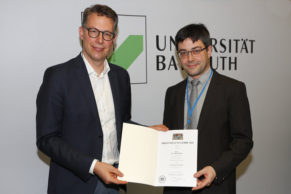 Zwei Männer in formeller Kleidung schauen in Richtung Betrachter und halten gemeinsam eine geöffente Urkunden-Mappe. Auf dem Dokument in der Mappe ist das Bayerische Staatswappen zu erkennen. (öffnet Vergrößerung des Bildes)