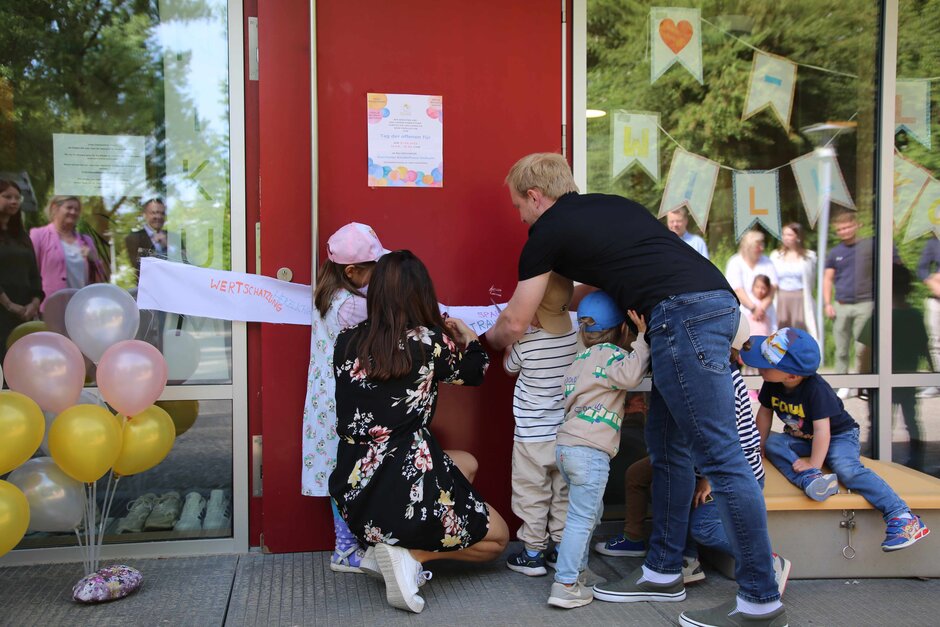 Halbnahe Aufnahme von einer Personengruppe vor einer roten Tür, links und rechts der Tür Glaswände, durch die man daran von innen befestigte Basteleien sieht. Die Personengruppe besteht aus einer Frau (hockend) und einem Mann (sich bückend) sowie fünf Kindern (alle etwa 3 Jahre alt). Alle haben dem Betrachter den Rücken zugewandt. Sie zwerschneiden gemeinsam ein weißes Band, das quer vor die Tür gespannt ist.