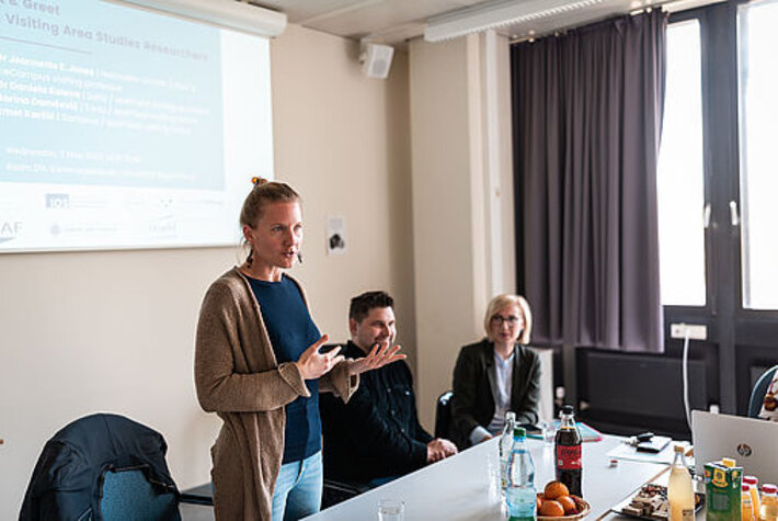 Eine Frau mit hochgesteckten Haaren und langen Ohrringen spricht in einem Konferenzraum. Neben ihr sitzen zwei Personen, die aufmerksam zuhören. (öffnet Vergrößerung des Bildes)