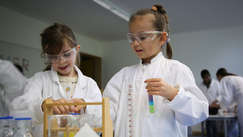 Zwei junge Mädchen in Kitteln und mit Schutzbrillen führen chemische Experimente durch (öffnet Vergrößerung des Bildes)