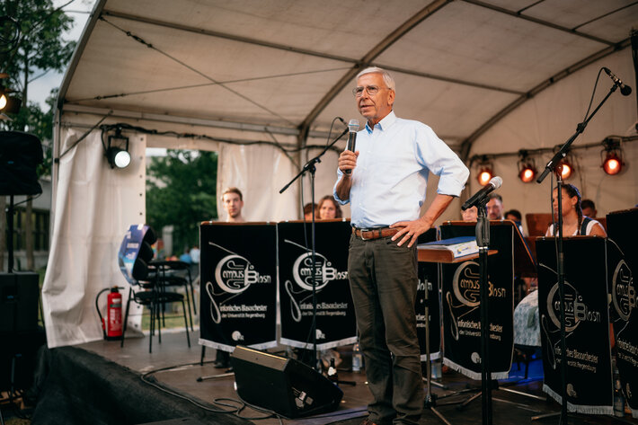 Begrüßung durch Universitätspräsident Prof. Dr. Udo Hebel. Ein Herr mit weißen Haaren und Brille steht auf der Bühne und spricht in ein Mikrofon. Hinter ihm sitzen Musikerinnen und Musiker des Musikensembles CampusBlosn. (öffnet Vergrößerung des Bildes)