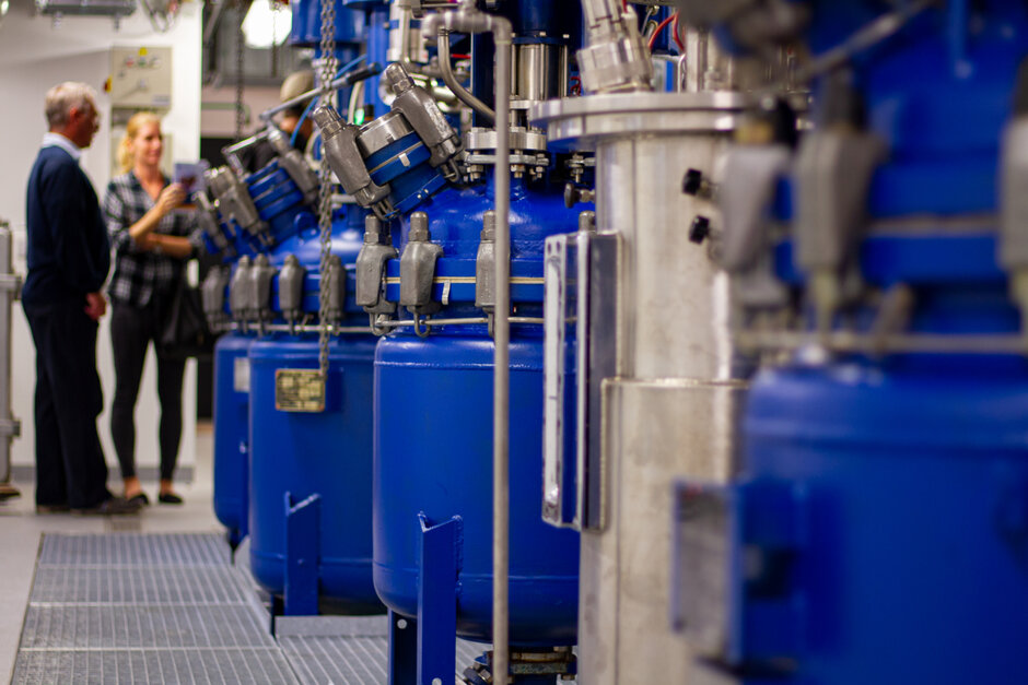 Eine Reihe großer blauer Tanks in einem Labor- bzw. Technikraum; im Hintergrund stehen zwei Personen im Gespräch.