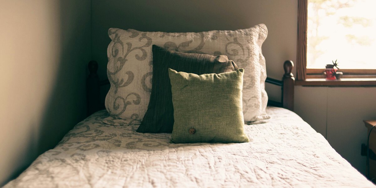 Zu sehen ist ein Einzelbett mit Decke und drei unterschiedlich großen Kissen. Rechts im Bild ist auch ein Fenster zu sehen.