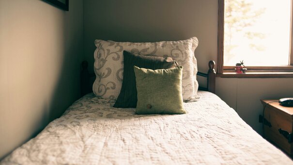 Zu sehen ist ein Einzelbett mit Decke und drei unterschiedlich großen Kissen. Rechts im Bild ist auch ein Fenster zu sehen.