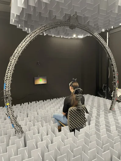 Eine Person sitzt auf einem Stuhl in einem Raum mit zahlreichen vertikal angeordneten weißen Paneelen. Die Person trägt ein Headset und hat Geräte am Rücken befestigt. Ein großer kreisförmiger Metallrahmen umgibt sie, und über ihr hängen weitere weiße Paneele von der Decke. Vor der Person befindet sich ein kleiner Bildschirm mit grafischer Darstellung. Der Raum wirkt wie eine Versuchsanordnung für ein Experiment mit sensorischer oder virtueller Realität.