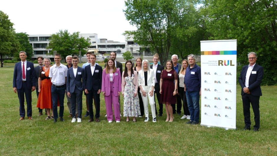 Ein Gruppenfoto mit mehreren Personen, die in feierlicher Kleidung neben einem Banner stehen, auf dem „RUL“ zu lesen ist. (öffnet Vergrößerung des Bildes)