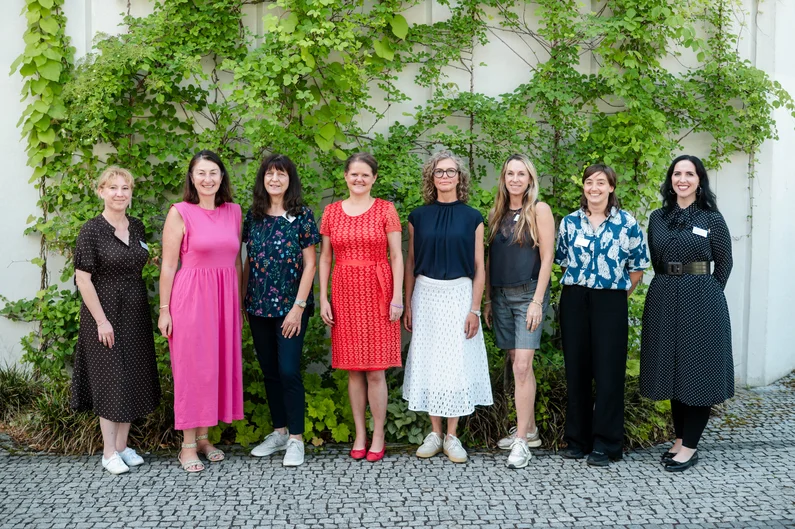 Acht Personen stehen für ein Gruppenfoto vor einer begrünten Wand mit Kletterpflanzen. Die Gruppe ist in einer Reihe auf einem gepflasterten Platz arrangiert. (öffnet Vergrößerung des Bildes)