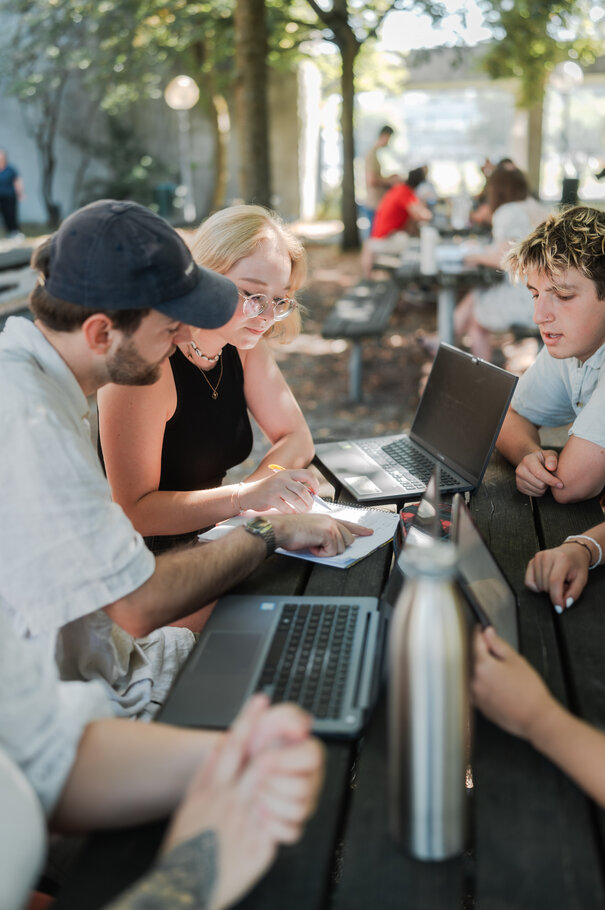 Studierende sitzen zusammen an einem Holztisch unter freiem Himmel. Auf dem Tisch stehen Laptops und eine Trinkflasche.