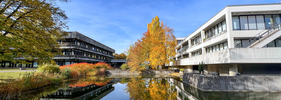 Blick über den Uni-Campus; im Vordergrund ein Teich, im Bildhintergrund Gebäude in Sichtbetonbauweise mit Flachdächern, Bäume und Sträucher mit herbstlich gefärbten Blättern