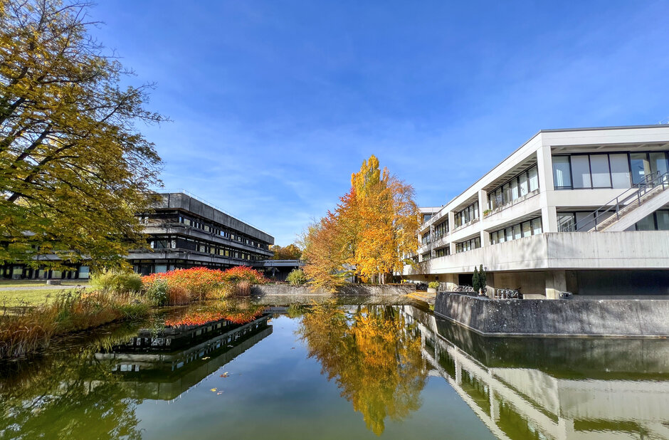 Blick über den Uni-Campus; im Vordergrund ein Teich, im Bildhintergrund Gebäude in Sichtbetonbauweise mit Flachdächern, Bäume und Sträucher mit herbstlich gefärbten Blättern