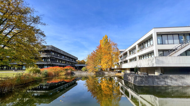 Blick über den Uni-Campus; im Vordergrund ein Teich, im Bildhintergrund Gebäude in Sichtbetonbauweise mit Flachdächern, Bäume und Sträucher mit herbstlich gefärbten Blättern