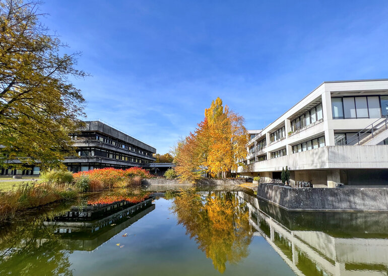 Blick über den Uni-Campus; im Vordergrund ein Teich, im Bildhintergrund Gebäude in Sichtbetonbauweise mit Flachdächern, Bäume und Sträucher mit herbstlich gefärbten Blättern