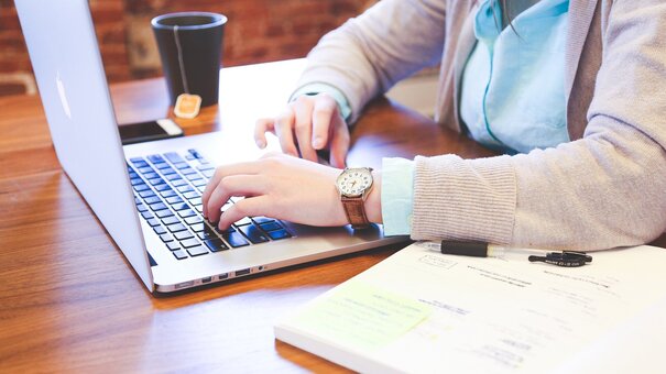 Eine Person sitzt am Schreitisch und tippt etwas auf einem Laptop. Daneben ein Notizblock, ein Smartphone und eine Tasse Tee. 