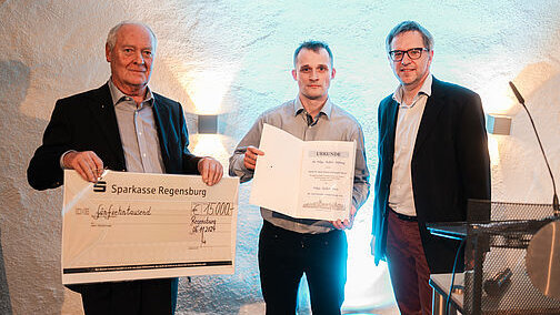 Drei Herren stehen nebeneinander. Der linke hält einen Dekoscheck in Händen, der mittlere hält eine Urkundenmappe hoch und zeigt die darin befindliche Urkunde.