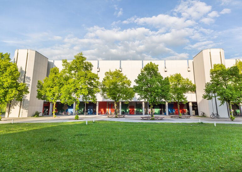 Blick über eine Wiese und Bäume auf den Gebäudeteil Philosophie/Theologie am Campus der Universität Regensburg
