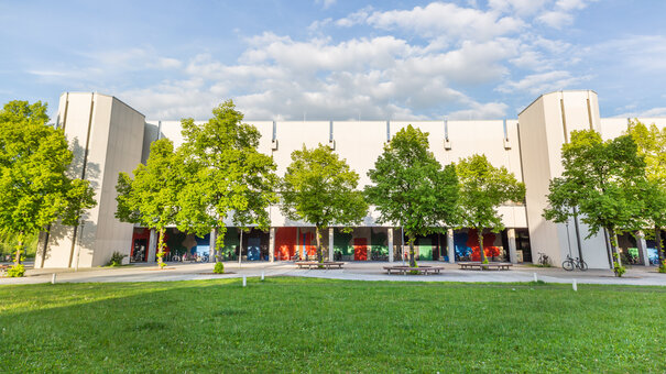 Blick über eine Wiese und Bäume auf den Gebäudeteil Philosophie/Theologie am Campus der Universität Regensburg