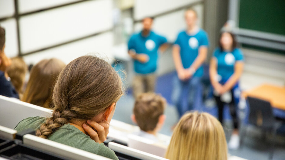 Ein Hörsaal in dem junge Personen von hinten zu sehen sind. Sie blicken auf drei Personen in türkis-farbenen T-Shirts, die am vorderen Ende des Hörsaals vor einer grünen Tafel stehen. Diese Personen sind nur verschwommen zu erkennen.