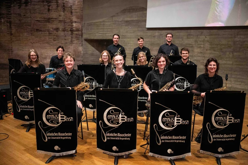 Mitglieder des Symphonischen Blasorchesters der Universität Regensburg sitzen mit ihren Instrumenten auf einer Bühne. Vor ihnen stehen schwarze Notenpulte mit dem Schriftzug „Campus Blosn“, im Hintergrund sind weitere Orchestermitglieder und eine Betonwand zu sehen. (öffnet Vergrößerung des Bildes)