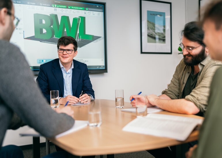 Vier Männer sitzen um einen Tisch, vor ihnen liegen Unterlagen, sie sprechen miteinander. Im Hintergrund ist eine elektronische Tafel zu sehen, auf der in großen Buchstaben "BWL" zu lesen ist.