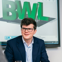 Vier Männer sitzen um einen Tisch, vor ihnen liegen Unterlagen, sie sprechen miteinander. Im Hintergrund ist eine elektronische Tafel zu sehen, auf der in großen Buchstaben "BWL" zu lesen ist.