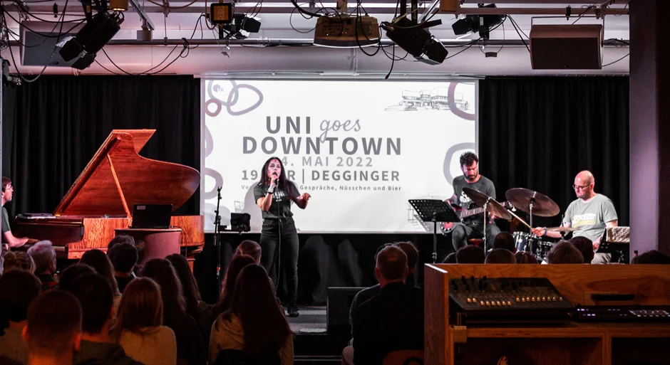 Vier Musiker*innen auf einer Bühne, eine Person sitzt an einem Flügel, eine Frau singt in ein Mikrofon, ein Mann spielt auf einer E-Gitarre und ein Mann spielt Schlagzeug. Im Hintergrund ist der Schriftzug "Uni goes Downtown" auf eine Leinwand projeziert.  (öffnet Vergrößerung des Bildes)