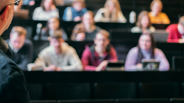 Ein älterer Mann steht in einem Hörsaal und erklärt einer Gruppe von Studierenden etwas.