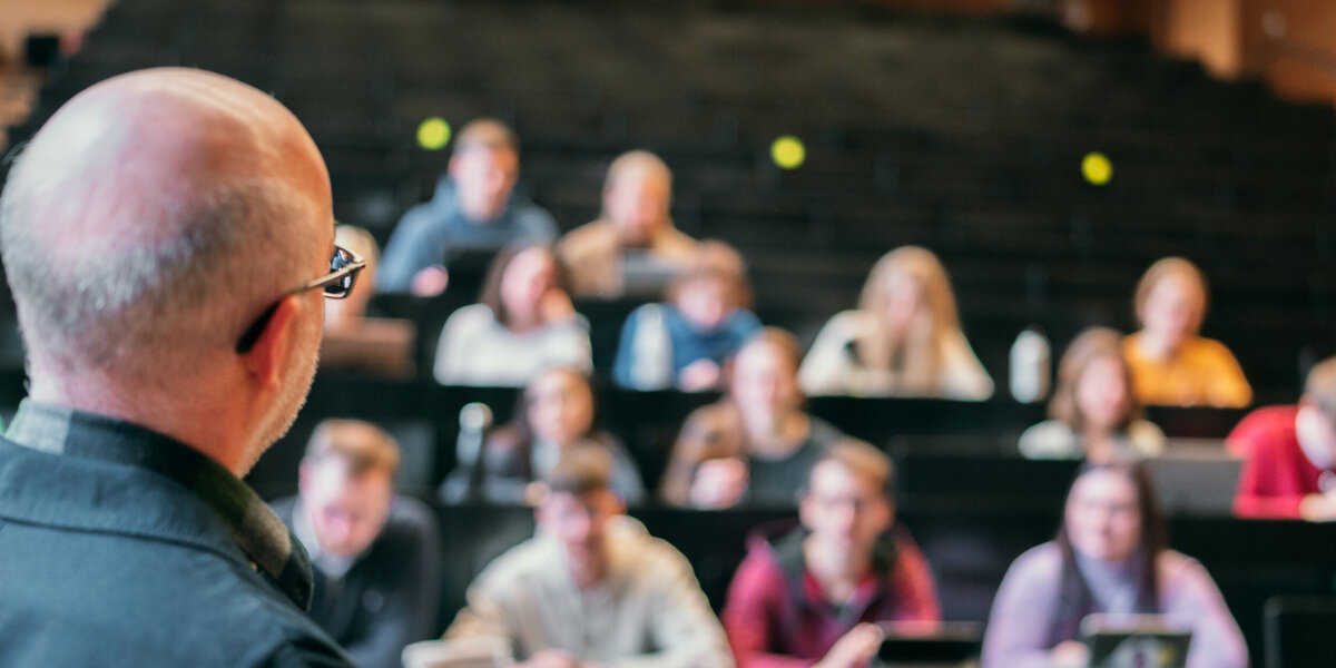 Ein älterer Mann steht in einem Hörsaal und erklärt einer Gruppe von Studierenden etwas.