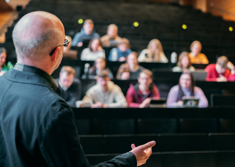Ein älterer Mann steht in einem Hörsaal und erklärt einer Gruppe von Studierenden etwas.