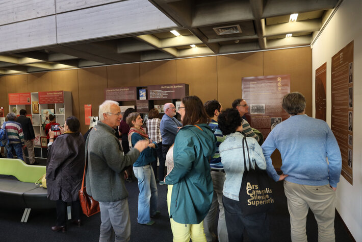 Mehrere Personen befinden sich in einer modernen Ausstellungsumgebung. Sie betrachten und diskutieren verschiedene Wandtafeln und Informationsdisplays. Der Raum ist gut beleuchtet, mit mehreren Vitrinen und Schautafeln mit Texten und Bildern. Einige Personen führen Gespräche, andere betrachten die Exponate aufmerksam. (öffnet Vergrößerung des Bildes)