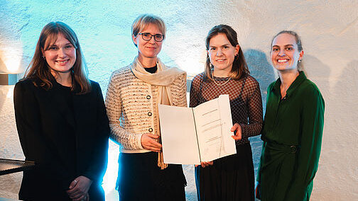 Vier Frauen stehen nebeneinander vor einer weißen Wand. Die mittleren beiden halten eine aufgeschlagene Urkundenmappe so hoch, dass die enthaltene Urkunde zu sehen ist.