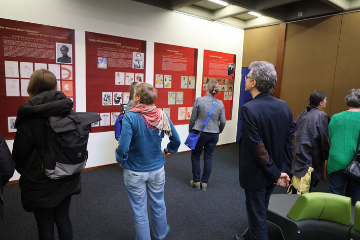 Mehrere Personen befinden sich in einer modernen Ausstellungsumgebung. Sie betrachten Wandtafeln mit Bildern und Informationstexten. Der Raum ist gut beleuchtet, es sind Sitzgelegenheiten zu sehen. Die Personen betrachten aufmerksam die Ausstellungsfläche. (öffnet Vergrößerung des Bildes)