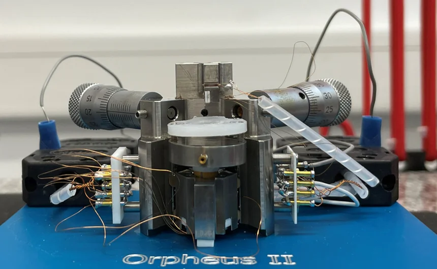 Das Foto zeigt ein wissenschaftliches Gerät auf einer blauen Unterlage mit der Aufschrift „Orpheus II“. Das Gerät besteht aus mehreren Metallkomponenten, darunter zwei Mikrometerschrauben an den Seiten und einer zentralen Einheit. An diese Einheit sind verschiedene Drähte und Schläuche angeschlossen. (öffnet Vergrößerung des Bildes)