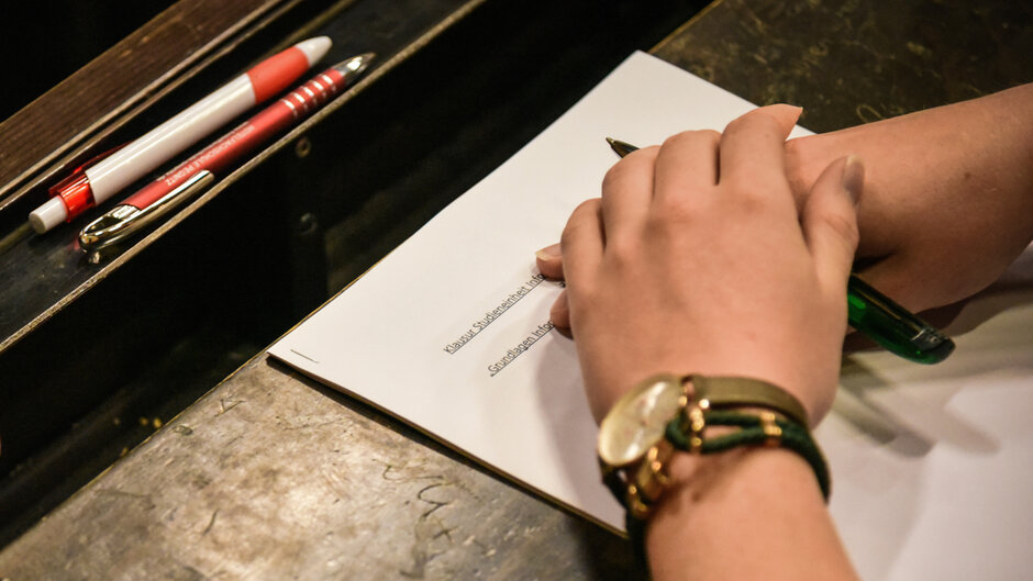 Auf einem Hörsaaltisch liegt ein Stapel DIN A4-Blätter; auf dem obersten Blatt ist das Wort "Klausur" zu sehen. Auf dem Blatt liegt ein Händepaar, darüber zwei Stifte.