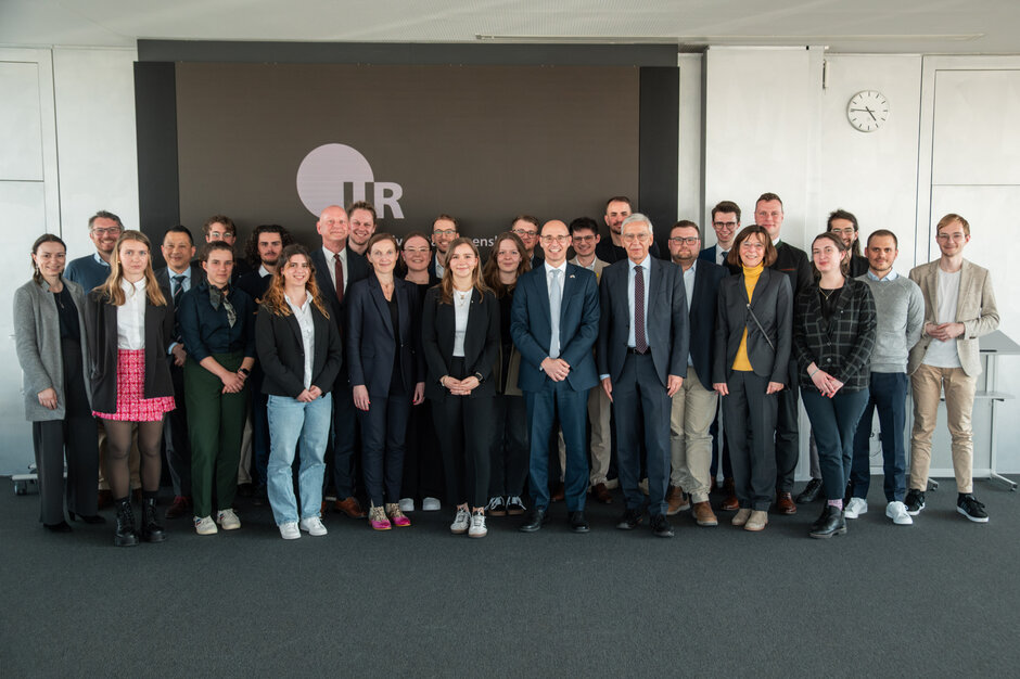 Ein Gruppenfoto mit über 25 Männern und Frauen unterschiedlichen Alters. Im Hintergrund ist ein großformatiger Bildschirm mit UR-Logo zu sehen. 