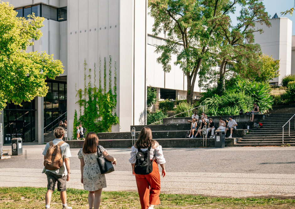 Drei Personen gehen auf ein Gebäude zu. Es ist Sommer, der Himmel ist blau, die Bäume sind grün, die Sonne scheint.