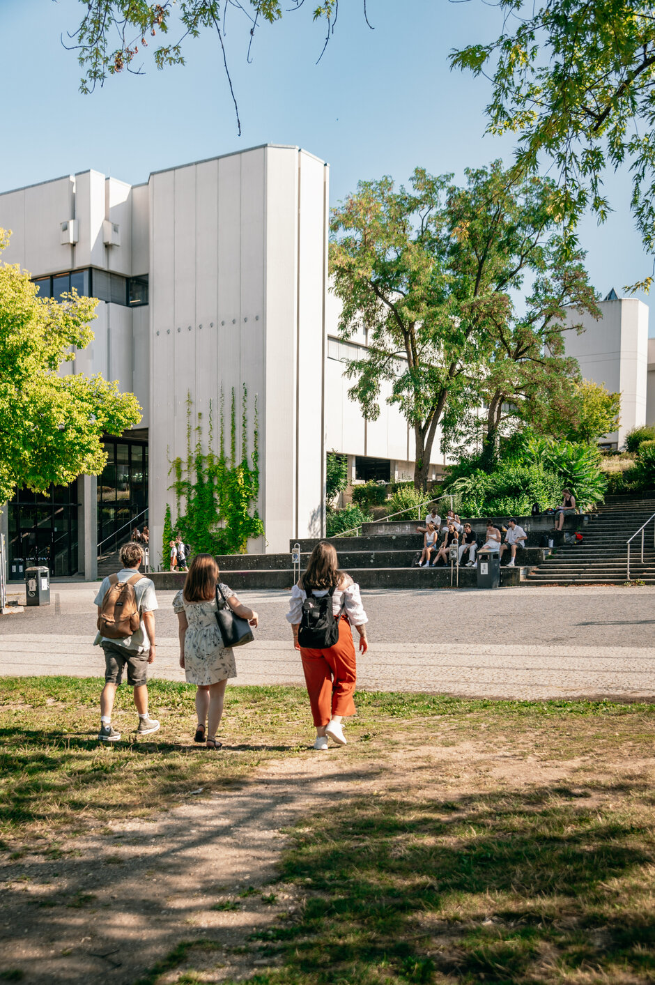 Drei Personen gehen auf ein Gebäude zu. Es ist Sommer, der Himmel ist blau, die Bäume sind grün, die Sonne scheint.