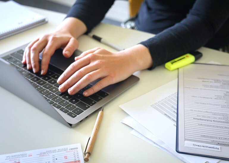 Preson, die auf einem Laptop etwas tippt. Daneben liegen Unterlagen und Stifte.