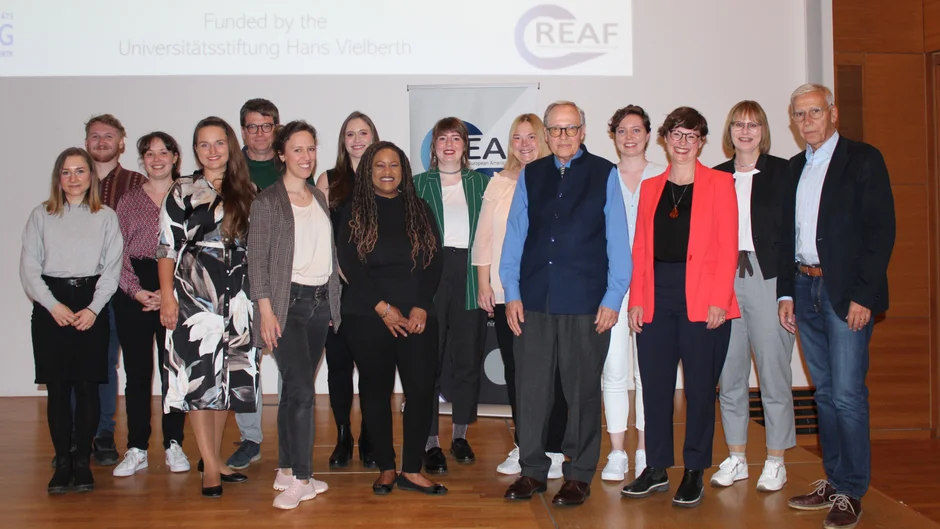 Gruppenbild mit ca. 15 Personen, die sich auf einer Bühne für ein Foto zusammengestellt haben.