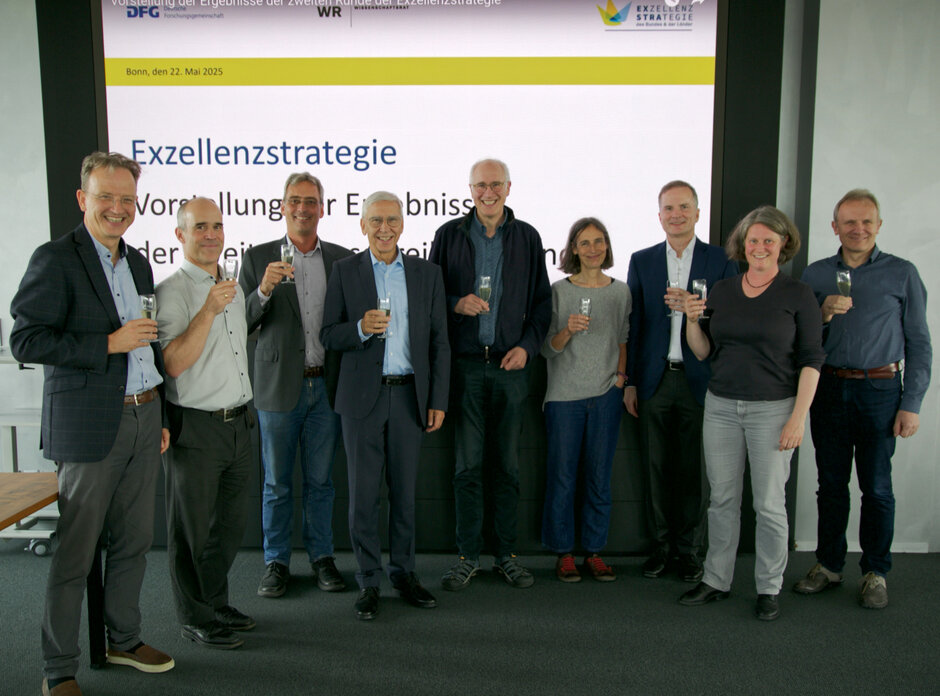 Eine Gruppe von Männern und Frauen in fröhlicher Stimmung steht zusammen mit Blick zur Kamera. Alle halten ein gefülltes Sektglas in der Hand.