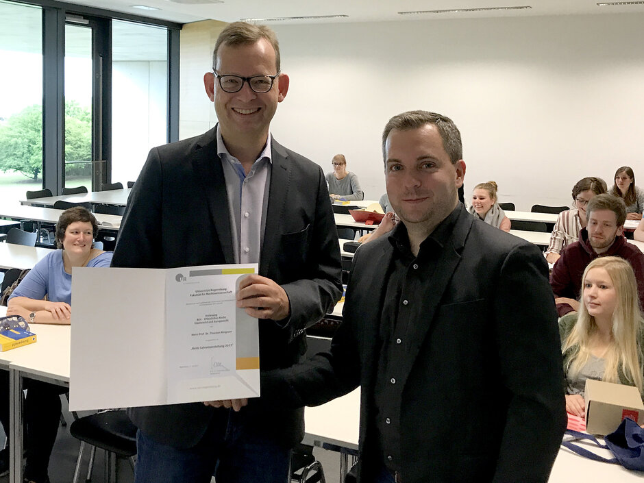 Prof. Kingreen mit der Urkunde in Händen und Herr Gailer stehen lächelnd in einem Seminarraum mit einigen Studierenden an ihren Sitzplätzen im Hintergrund. (öffnet Vergrößerung des Bildes)