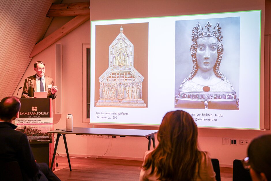 Mann mit Brille, gestikulierend, am Vortragspult; im Vordergrund Publikum.