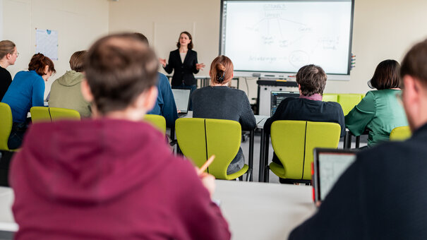 Zu sehen ist eine Dozentin, die neben einem Whiteboard steht und Studierende unterrichtet. Die Studierenden sitzen mit dem Rücken zum Betrachter. 