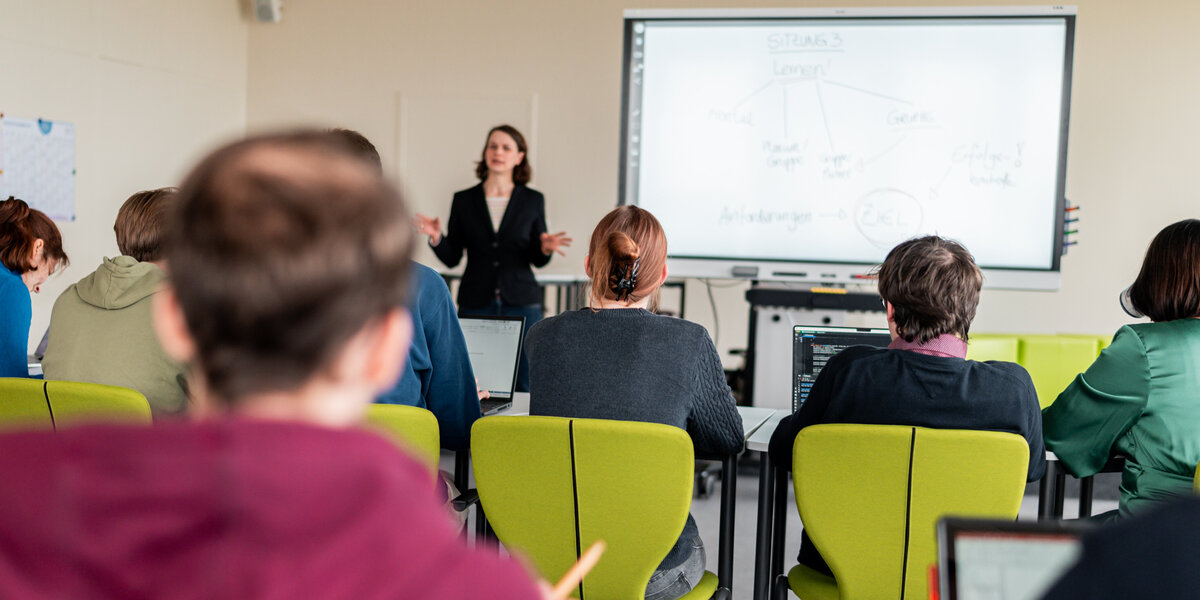 Zu sehen ist eine Dozentin, die neben einem Whiteboard steht und Studierende unterrichtet. Die Studierenden sitzen mit dem Rücken zum Betrachter. 