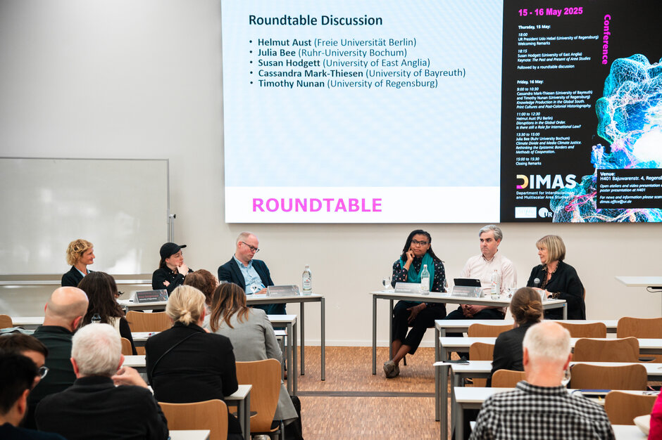 Die sechs Teilnehmerinnen und Teilnehmer des Roundtable sitzen im Halbkreis mit dem Gesicht zur Kamera. Im Bildvordergrund sind Hinterköpfe des Publikums zu sehen. (öffnet Vergrößerung des Bildes)