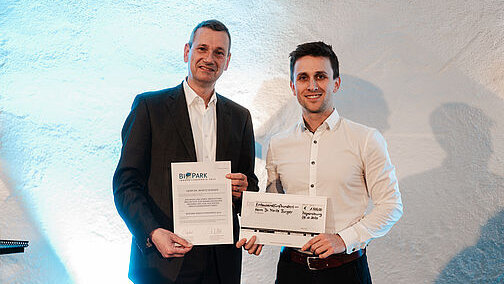 Zwei Männer halten eine Urkunde und einen Dekoscheck in die Kamera. Der eine trägt einen dunklen Anzug, der andere eine dunkle Hose und ein weißes Hemd.